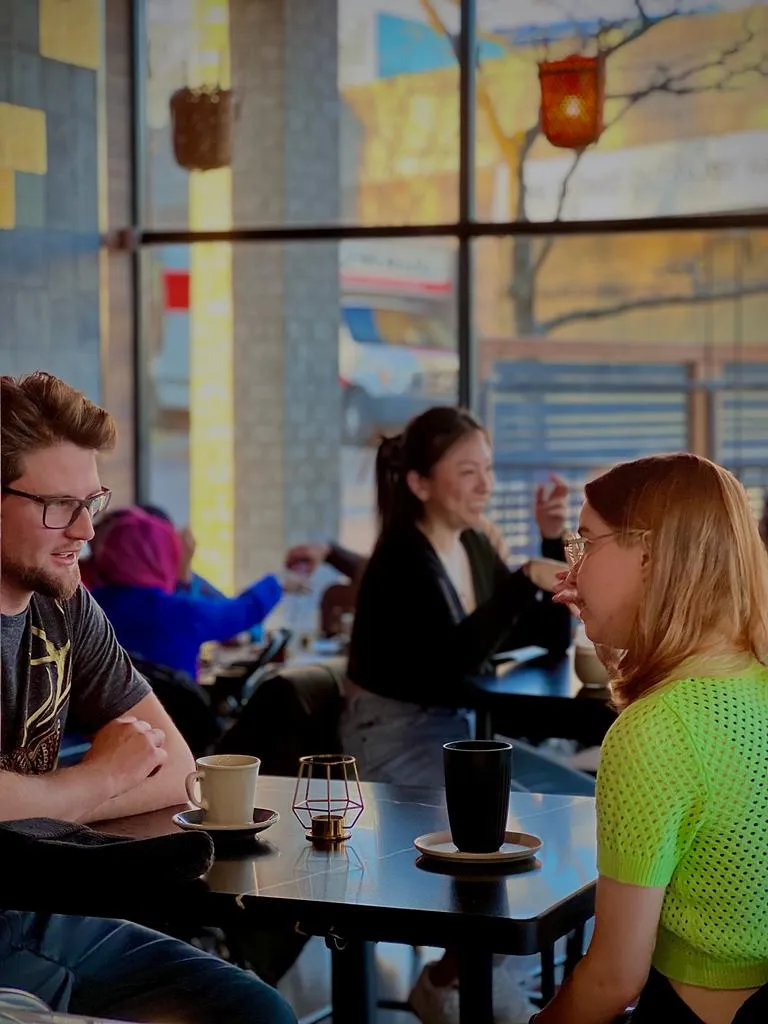 Two people sitting at a cafe table, drinking coffee and talking inside a bright coffee shop.
