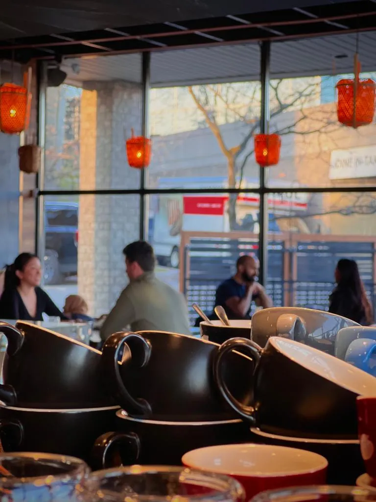 Stacked coffee cups in a cafe with four people conversing near large windows and orange lights.