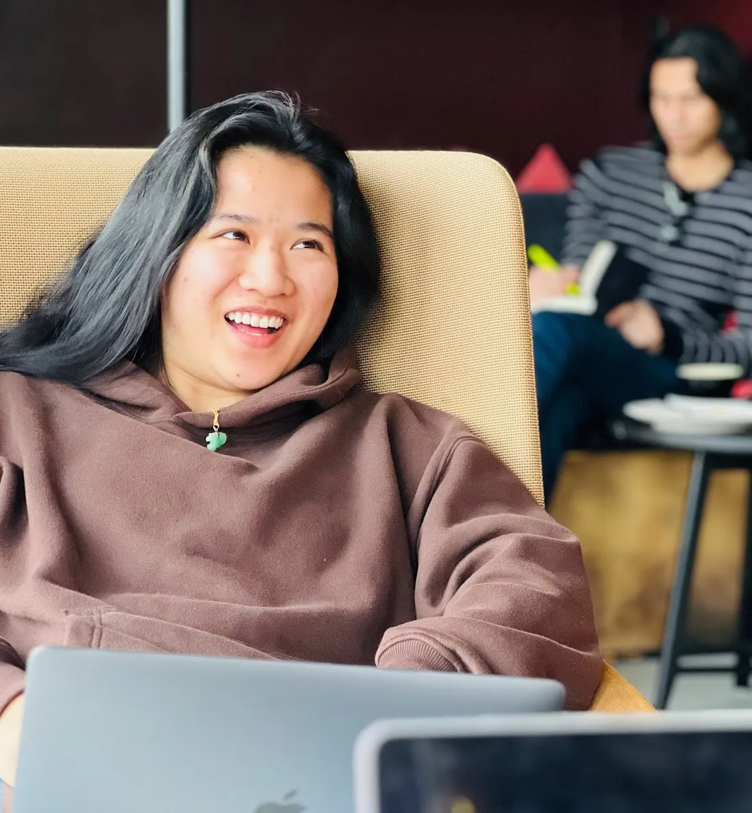 Smiling woman with long black hair wearing brown hoodie sitting with a laptop, another person writing in background.